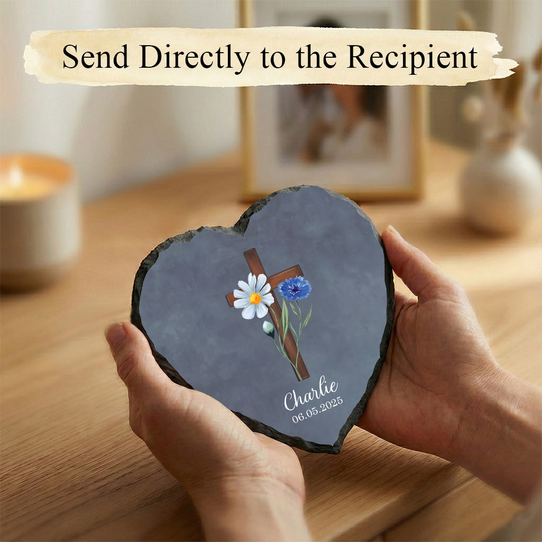 Heart-shaped slate with a cross and flowers design held by a hand on a wooden surface.